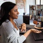 A woman takes a conference call in her home office. Two computer screens are filled with the headshots of participants.