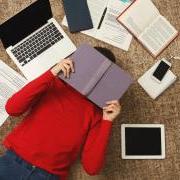 Student lying on the floor with a book over their face surrounded by papers, a laptop, phone and textbooks.